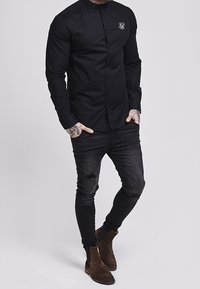 Zwart overhemd met knopen en een opstaande kraag met een geborduurd logo op de linkerborst. Draagt zwart versleten skinny jeans en bruine laarzen.