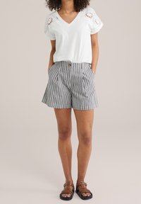 Donna che indossa una blusa bianca con maniche a occhiello, pantaloni corti a righe bianche e nere a vita alta e sandali marroni con fibbia, in piedi con le mani in tasca.