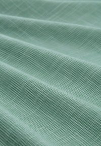 Světlé zelené texturované látky s mřížkovým vzorem; záhyby vytvářejí jemné stíny, což zdůrazňuje tkaní a povrchové detaily materiálu.