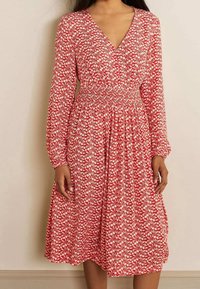 Vestido rojo con un escote en V profundo, mangas largas y cintura fruncida. Presenta un patrón floral blanco en una tela ligera. La falda acampanada llega hasta la mitad de la pantorrilla.