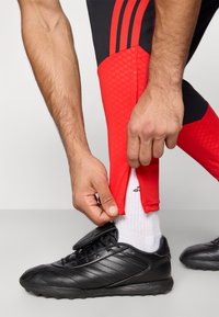 Sapatilha atlética preta com detalhes texturizados, combinada com calças justas vermelhas e pretas, mostrando uma mão a ajustar a bainha acima de uma meia branca.