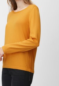 Pull jaune avec un col rond, des manches longues et des fentes sur le côté. Fabriqué en tissu doux avec une texture lisse. Détail du logo en petit au bas.