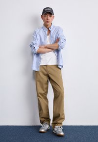 Cămașă cu dungi albastre și albe, purtată peste un tricou alb, asociată cu pantaloni bej și adidași argintii; poartă o șapcă gri de baseball.
