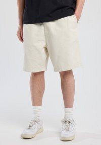 Pantalones cortos de algodón beige con un ajuste relajado, hasta la rodilla, con un dobladillo recto y bolsillos laterales, combinados con zapatillas blancas y calcetines.