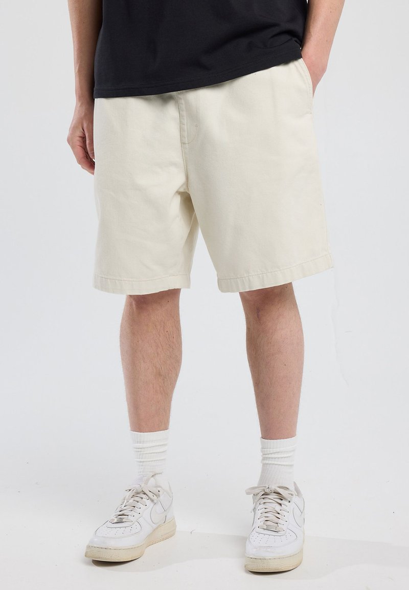 Pantalones cortos de algodón beige con un ajuste relajado, hasta la rodilla, con un dobladillo recto y bolsillos laterales, combinados con zapatillas blancas y calcetines.