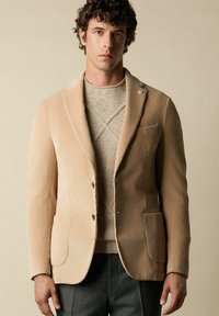 Beige korduroy sako s klopami, se dvěma předními kapsami a dvěma knoflíky. Nosit se na texturovaném béžovém svetru.