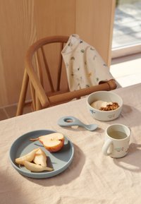 Piatto blu in silicone con fette di pera, ciotola bianca con granola, cucchiaio blu e tazza con motivi giocosi sopra una leggera tovaglia di lino.