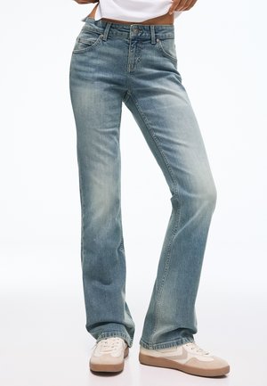 Personne portant un jean bleu clair droit et des baskets blanches avec des accents beiges sur fond blanc.