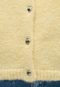 Gul strikket cardigan med myk tekstur, med sølvfargede metallknapper og ribbet kant, sammen med denimjeans.