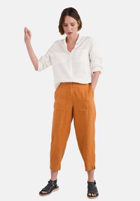 White linen button-up shirt with two chest pockets, paired with orange cropped pants featuring two front pockets. Black strappy sandals.
