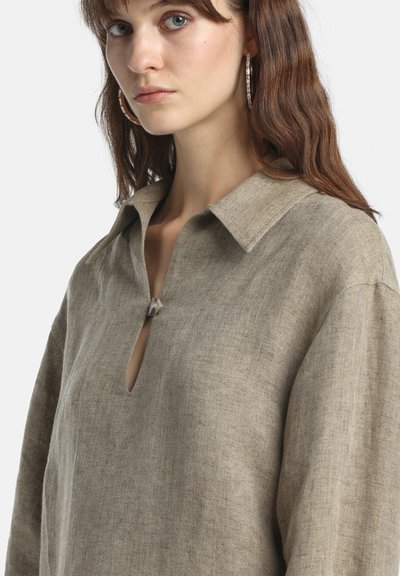 Fiatal nő világosbarna hajjal, nagy karika fülbevalókkal, laza, bézs lenvászon felsőben, széles gallérral és elöl gombokkal.