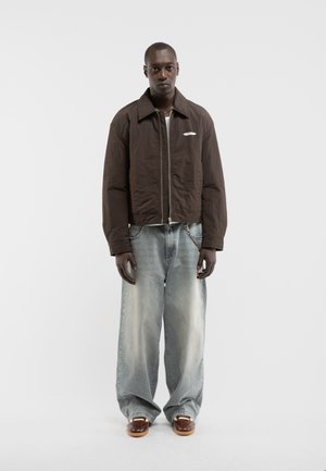 Homme debout portant une veste marron zippée, un jean large délavé avec une chaîne, et des chaussures marron sur un fond blanc uni.