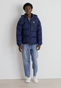 Giacca imbottita blu navy con colletto alto, zip, tasca sul petto e dettaglio del logo, abbinata a jeans in denim azzurri e sneakers bianche.