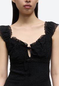 Kvinne iført en svart bluse uten ermer med blonder, rysjekanter på stroppene og en knute foran ved halsutskjæringen, som viser skuldrene og øvre del av brystet.