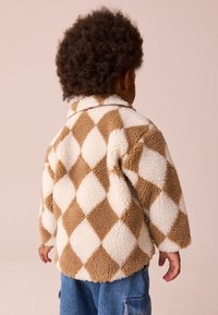 Braun-weiße Fleecejacke mit Rautenmuster, Kragen und langen Ärmeln, mit einer weichen, strukturierten Oberfläche. Sichtbare Jeanshose.