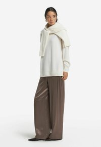 Weißer übergroßer Pullover mit geripptem Saum, kombiniert mit weit geschnittenen braunen Satin-Hosen. Ein passender weißer Schal fällt über den Pullover.