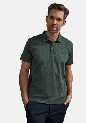 Mężczyzna ubrany w ciemnozieloną, wzorzystą koszulkę polo i ciemne spodnie, z rękami w kieszeniach, na jasnym, jednolitym tle.