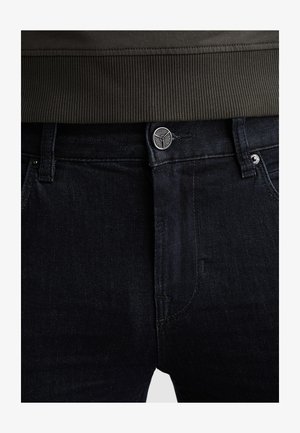 Schwarze Jeans aus Denim mit Knopfverschluss an der Taille und Gürtelschlaufen, kombiniert mit einem dunklen, gerippten Pullover, der leicht oberhalb des Bunds in die Hose gesteckt ist.