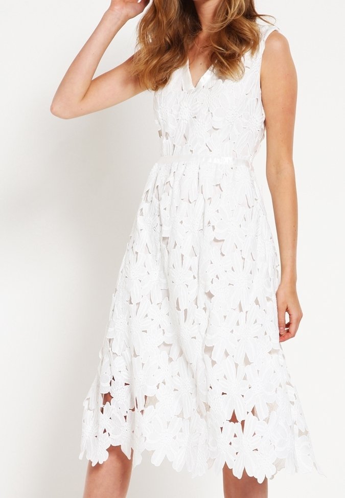 Femme portant une robe blanche en dentelle sans manches avec des motifs floraux découpés et un col en V, posant avec une main près de sa tête.