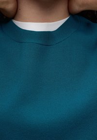 Turkusowy sweter z dzianiny z ribbowanym okrągłym dekoltem. Miękka tekstura i średnia waga, z białą wewnętrzną taśmą kołnierzyka widoczną przy dekolcie.