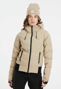 Beige Daunenjacke mit hohem Kragen, Reißverschluss vorne, schwarzen Akzenten und verstellbaren Kordeln. Kombiniert mit einer beige gestrickten Mütze.