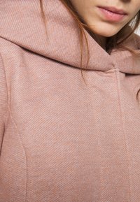 Zachtroze, gestructureerde stoffen hoodie met een brede, gevouwen capuchon en subtiele verticale naaddetails. Zacht en comfortabel uiterlijk, geschikt voor casual draagstijl.