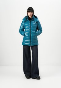 Vrouw in een turquoise pufferjack met capuchon, zwarte wijde broek en zwarte schoenen, staand tegen een witte achtergrond met handen in de zakken.