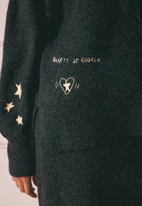 Schwarzer Strickpullover mit gerippten Bündchen und Saum. Mit goldener Stickerei des Textes "ROCKETT ST GEORGE" und dekorativen Sternmotiven.