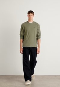 Olijfgroene geribde langemouwen shirt met een ronde halslijn, gecombineerd met losse zwarte broeken en beige sneakers op een neutrale achtergrond.