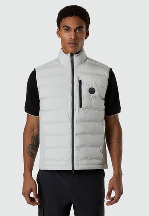 Uomo che indossa un gilet trapuntato grigio chiaro senza maniche con cerniera frontale e tasca sul petto sopra una maglietta nera a maniche corte, in piedi davanti a uno sfondo semplice.