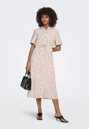 Femme en robe midi beige à pois noirs, tenant un petit sac noir, portant des sandales noires à talons ouverts, debout sur fond blanc.