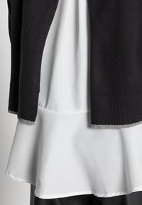 Black knit cardigan with silver trim overlapped on a white blouse, featuring a flared bottom hem and smooth texture. Black bottom visible.