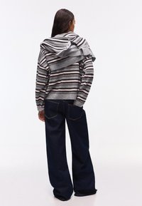 Donna in piedi di spalle, indossa un maglione a righe grigie e rosa e jeans larghi scuri, maglione appoggiato sulle spalle.