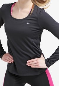 Musta pitkähihainen urheiluaita, jossa on pyöreä kaula-aukko ja hopea Nike-logo rinnassa, yhdistettynä tiukkoihin mustiin ja vaaleanpunaisiin housuihin.
