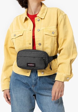 Osoba nosząca żółtą kurtkę, czerwoną koszulę, niebieskie dżinsy i ciemnoszarą saszetkę Eastpak na biodrach na jednolitym tle.