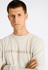 Sudadera gris claro con el texto "COPENHAGUE LINDBERGH" en beige contrastante. Tela suave con cuello redondo y corte relajado.