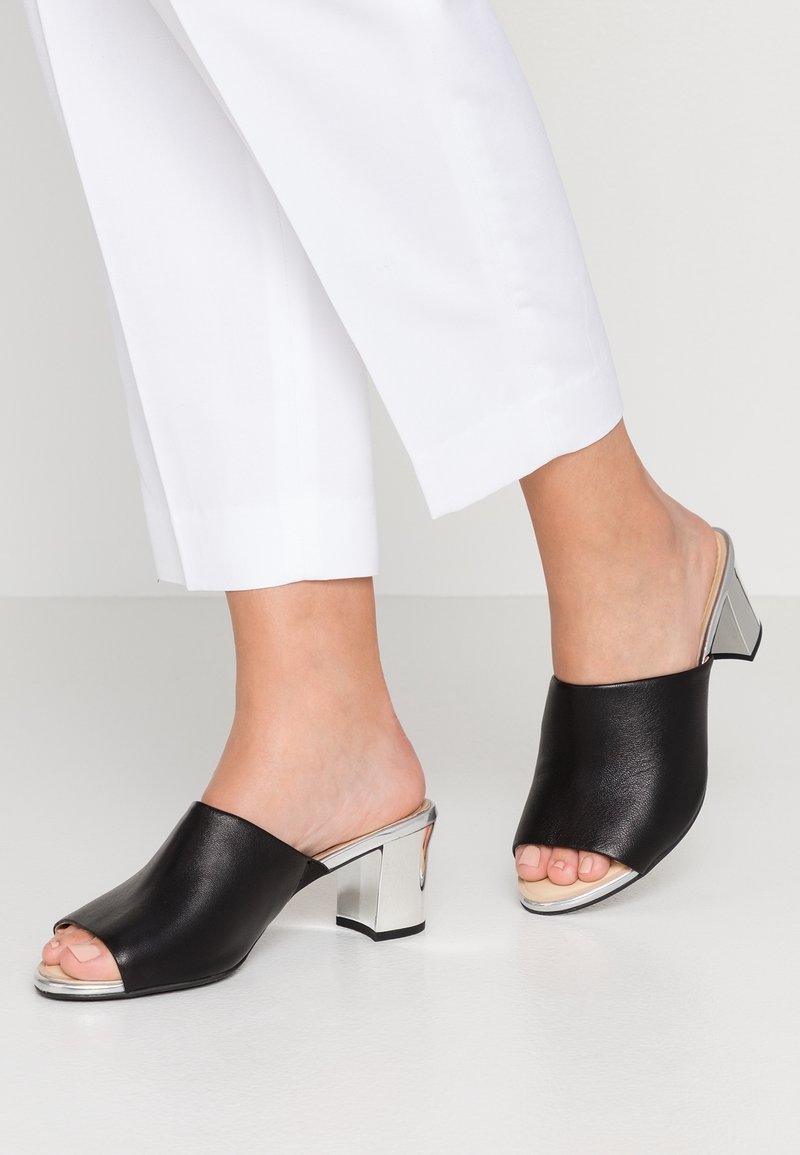 Svarta läderöppna mules med metallic silver blockklack. Har en slät textur och en minimalistisk design utan dekorationer.
