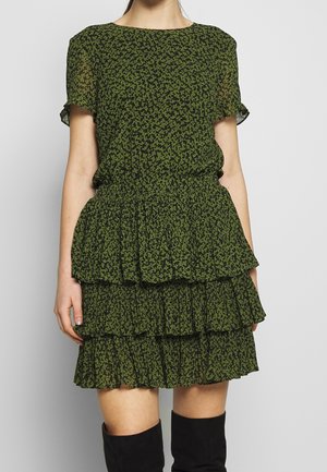 Femme portant une robe à volants à manches courtes avec un imprimé floral vert et noir, associée à des bottes noires montantes jusqu'aux genoux, sur un fond clair uni.