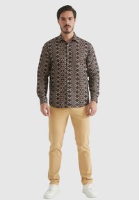Chemise à boutons dans un brun foncé avec des motifs géométriques en blanc et rouge. Associée à un pantalon beige clair et des baskets blanches.