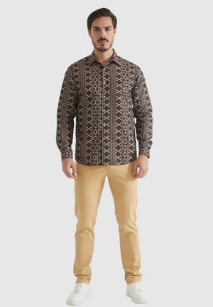 Chemise à boutons dans un brun foncé avec des motifs géométriques en blanc et rouge. Associée à un pantalon beige clair et des baskets blanches.