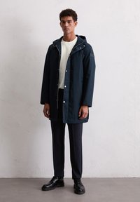 Jonge man in een marineblauwe hoodie met een witte sweatshirt, donkere broek en zwarte veterschoenen, staand tegen een eenvoudige witte muur.