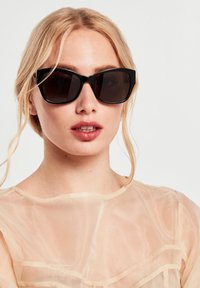 Gafas de sol negras de gran tamaño con lentes oscuros. Montura de plástico suave con bordes angulosos. Llevadas con una blusa semitransparente de color beige claro.