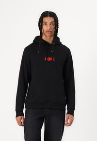 Black hoodie made of cotton with a front pocket and drawstring hood. Features a red logo appliqué on the chest. Casual fit and texture.