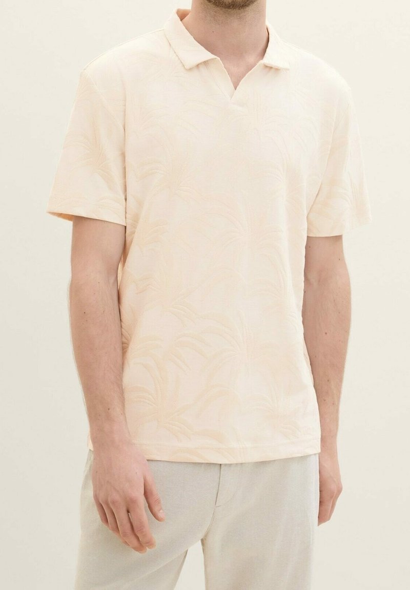Vaalea persikanvärinen polo-paita lyhyillä hihoilla, jossa on hienovarainen palmulehtikuosi, pehmeä tekstuuri ja klassinen kaulusuunnittelu.