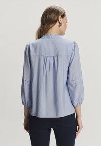 Blusa azzurra chiara con colletto alto, dettaglio arricciato sul retro e maniche a trequarti. Realizzata in un tessuto morbido e leggermente lucido.