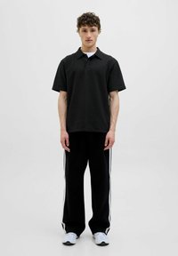 Polo noir à manches courtes avec un motif texturé, associé à un pantalon de survêtement noir avec des rayures blanches sur les côtés et des chaussures de sport.