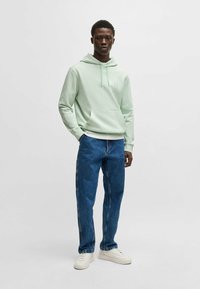 Mint green hoodie with kangaroo pocket, drawstring hood, and a logo patch. Paired with blue denim jeans and white sneakers.