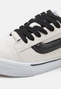 Gros plan d'une basket beige avec des lacets noirs, motif en vague noire sur le côté, semelle en caoutchouc blanche avec bout texturé.