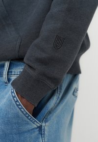 Personne portant un sweat-shirt gris foncé avec un logo brodé sur la manche, la main dans la poche d'un jean en denim bleu clair.