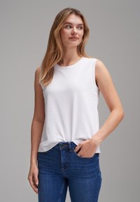 White sleeveless top made of soft cotton, featuring a round neckline, paired with blue jeans, displaying a classic fit and subtle stitching details.